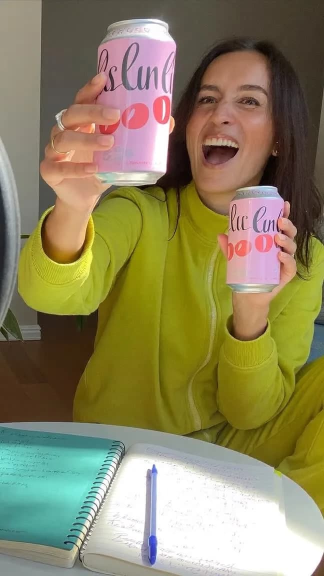 Laughing creator in a lime sweater holding up two pink cans