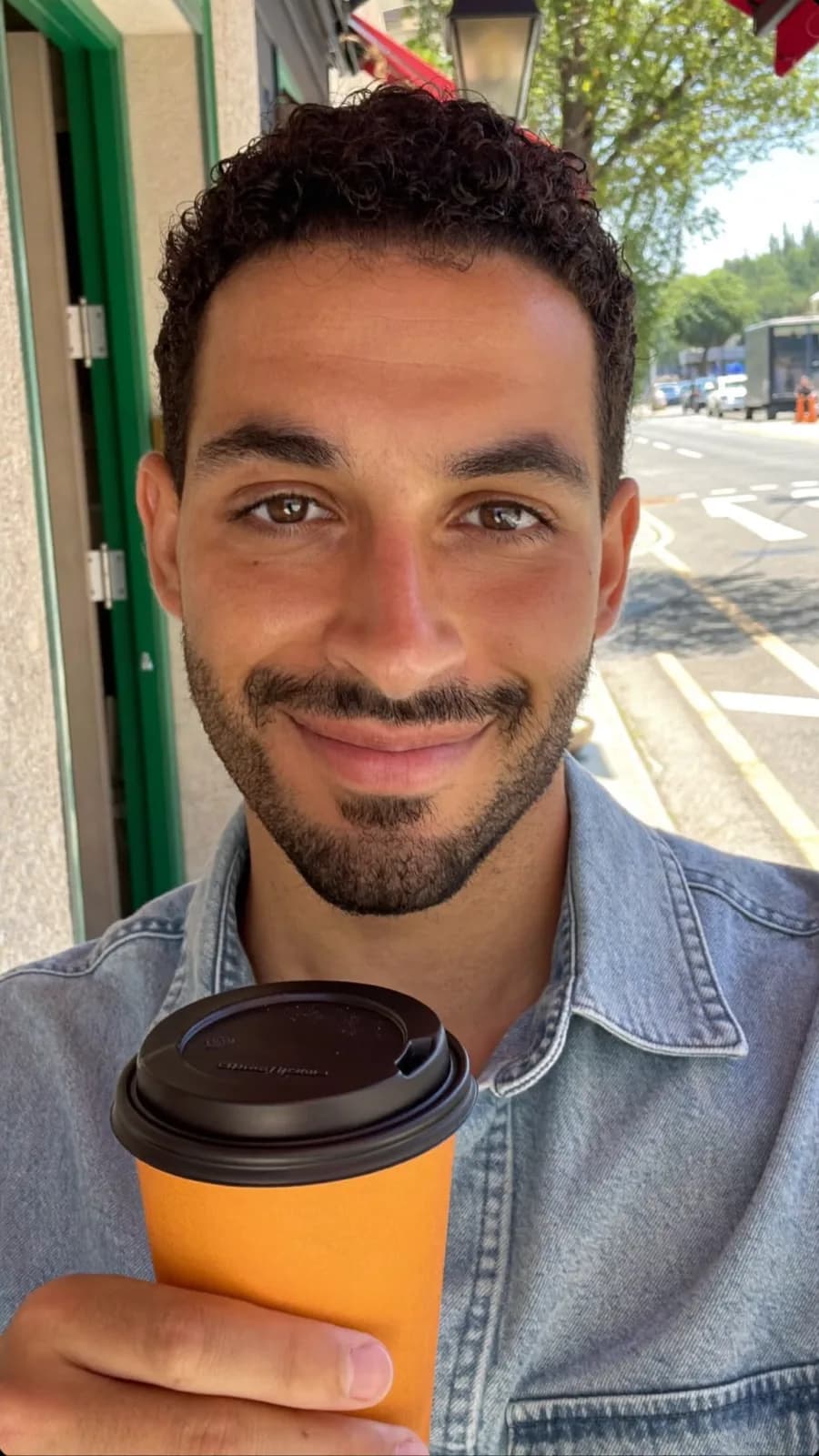 Creator in a denim shirt holding an orange takeaway coffee cup on a city street