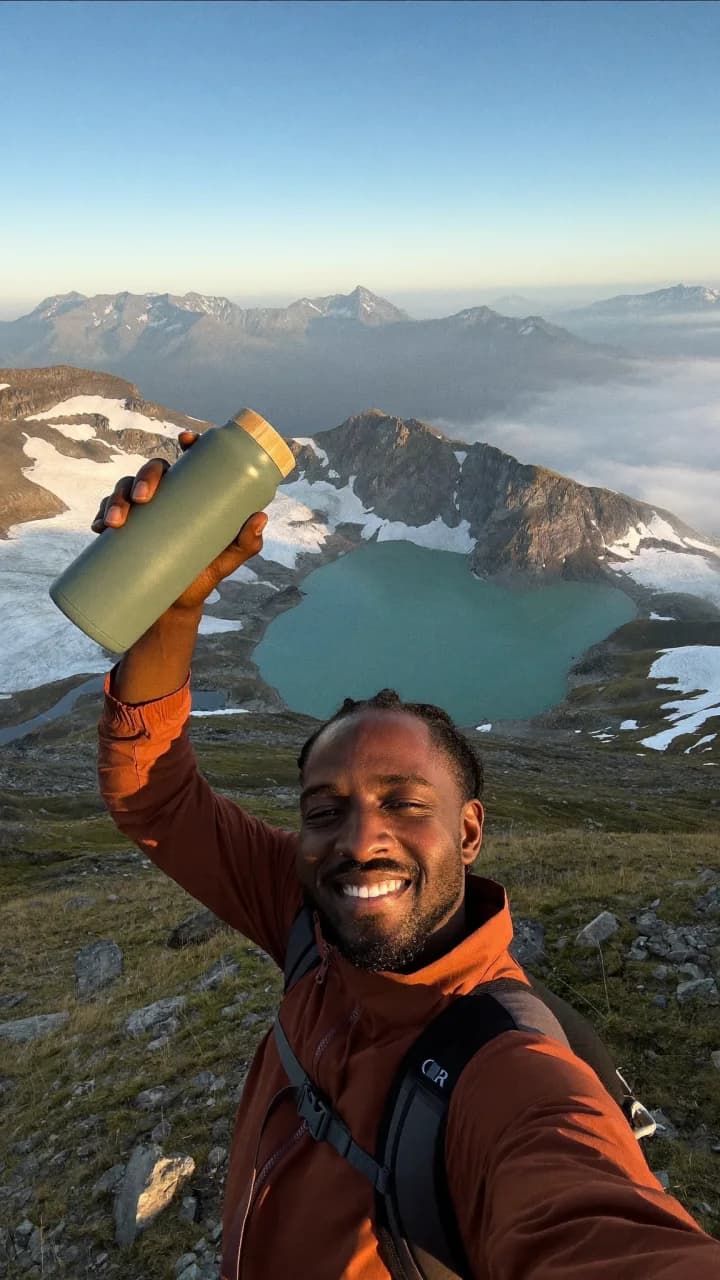 Hiker on a mountain summit raising a sage water bottle above a glacier lake