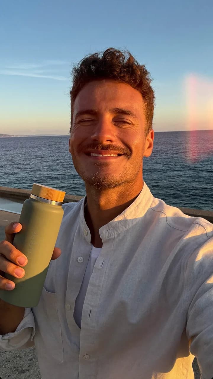 Smiling man in an off-white linen shirt holding a sage bottle in a seaside selfie at golden hour
