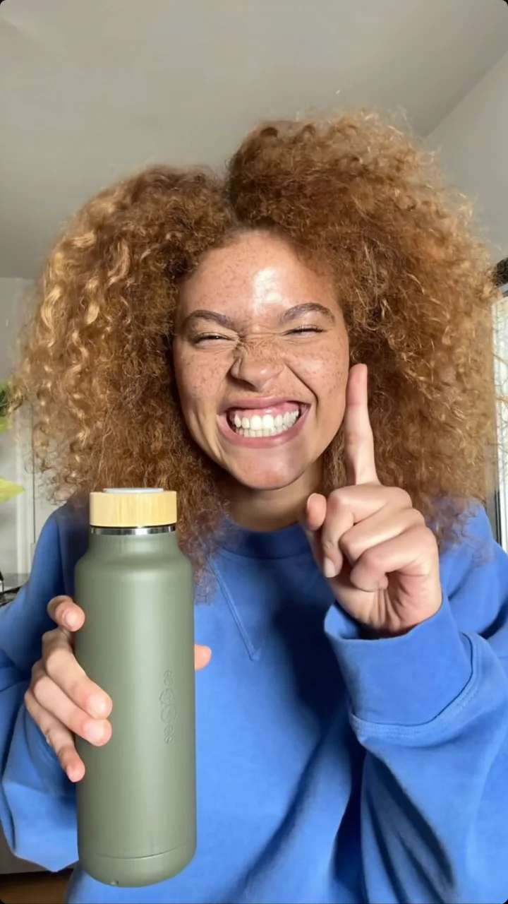 Laughing creator with curly hair in a blue sweatshirt holding a sage bottle, one finger raised