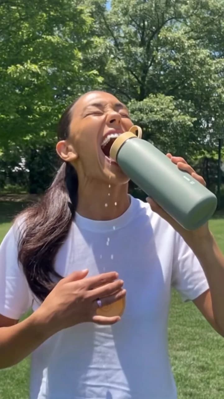 Woman drinking from a sage water bottle in a sunny park