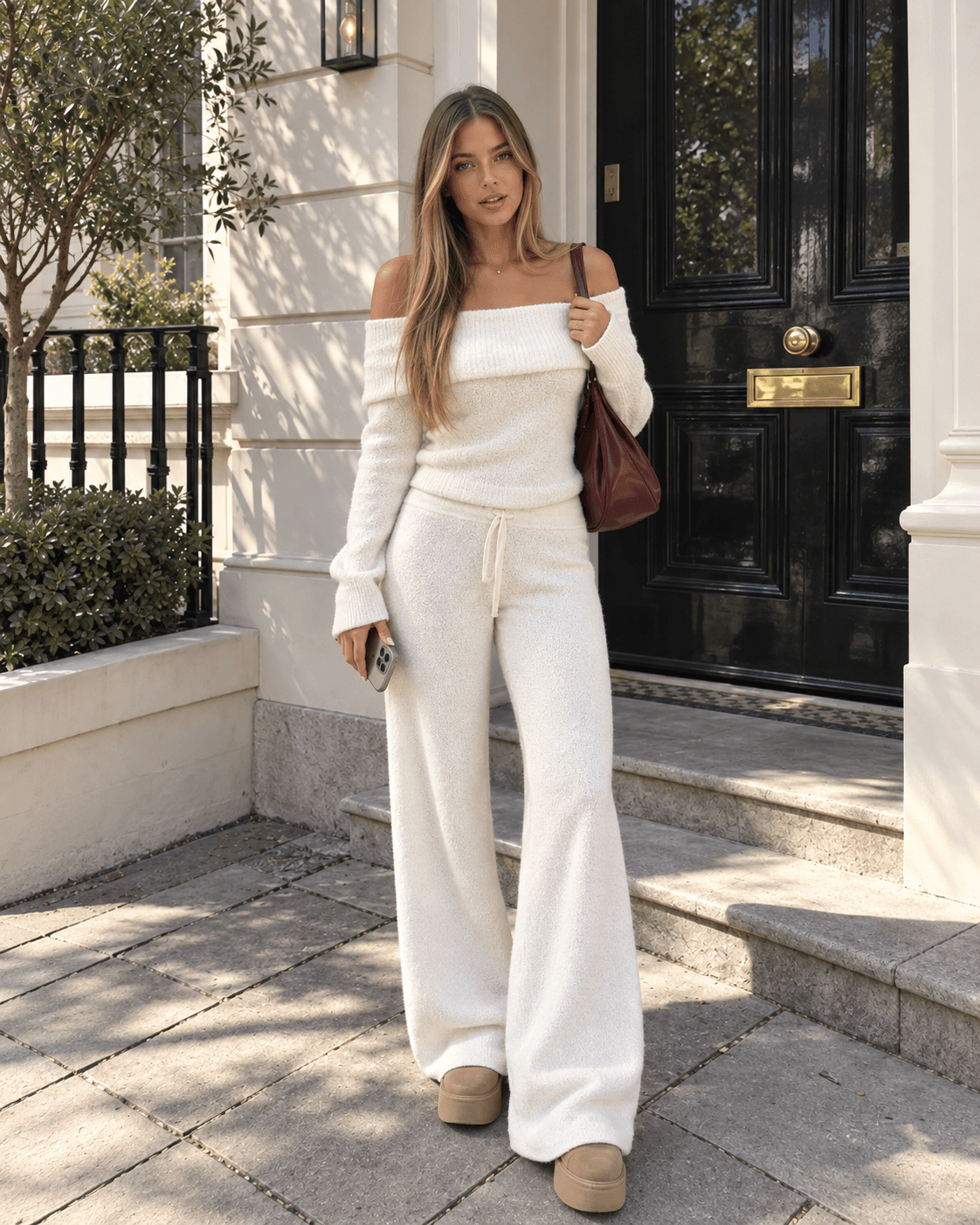 Woman in an off-shoulder cream knit and wide trousers with a burgundy bag before a black townhouse door