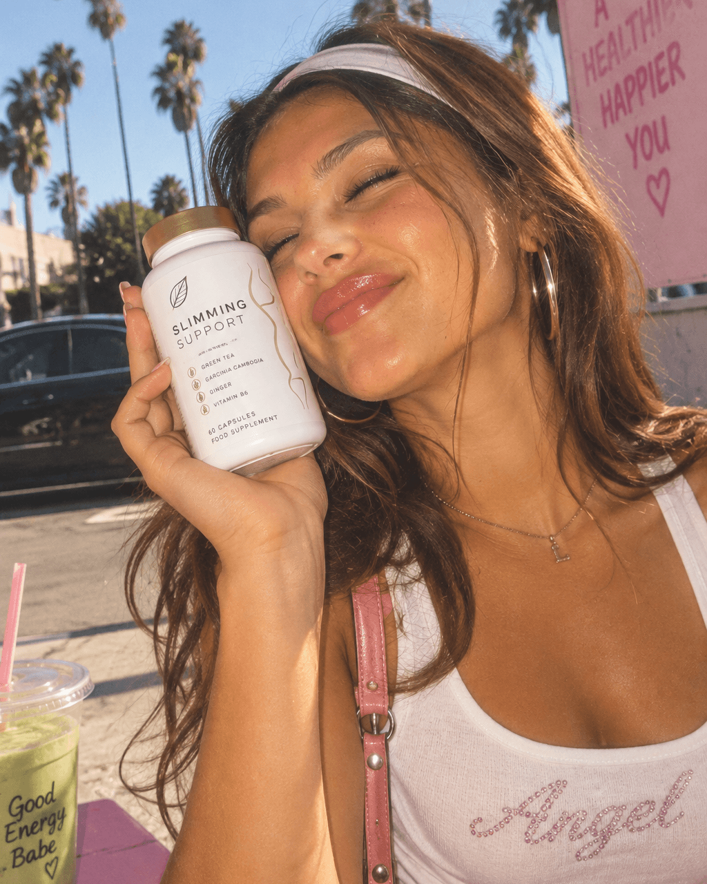 Smiling woman pressing a white supplement jar to her cheek on a sunny street