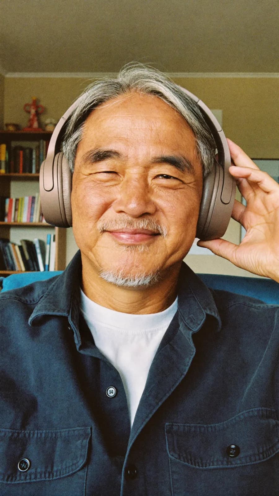 Man with long grey hair in a denim shirt wearing over-ear headphones in front of bookshelves