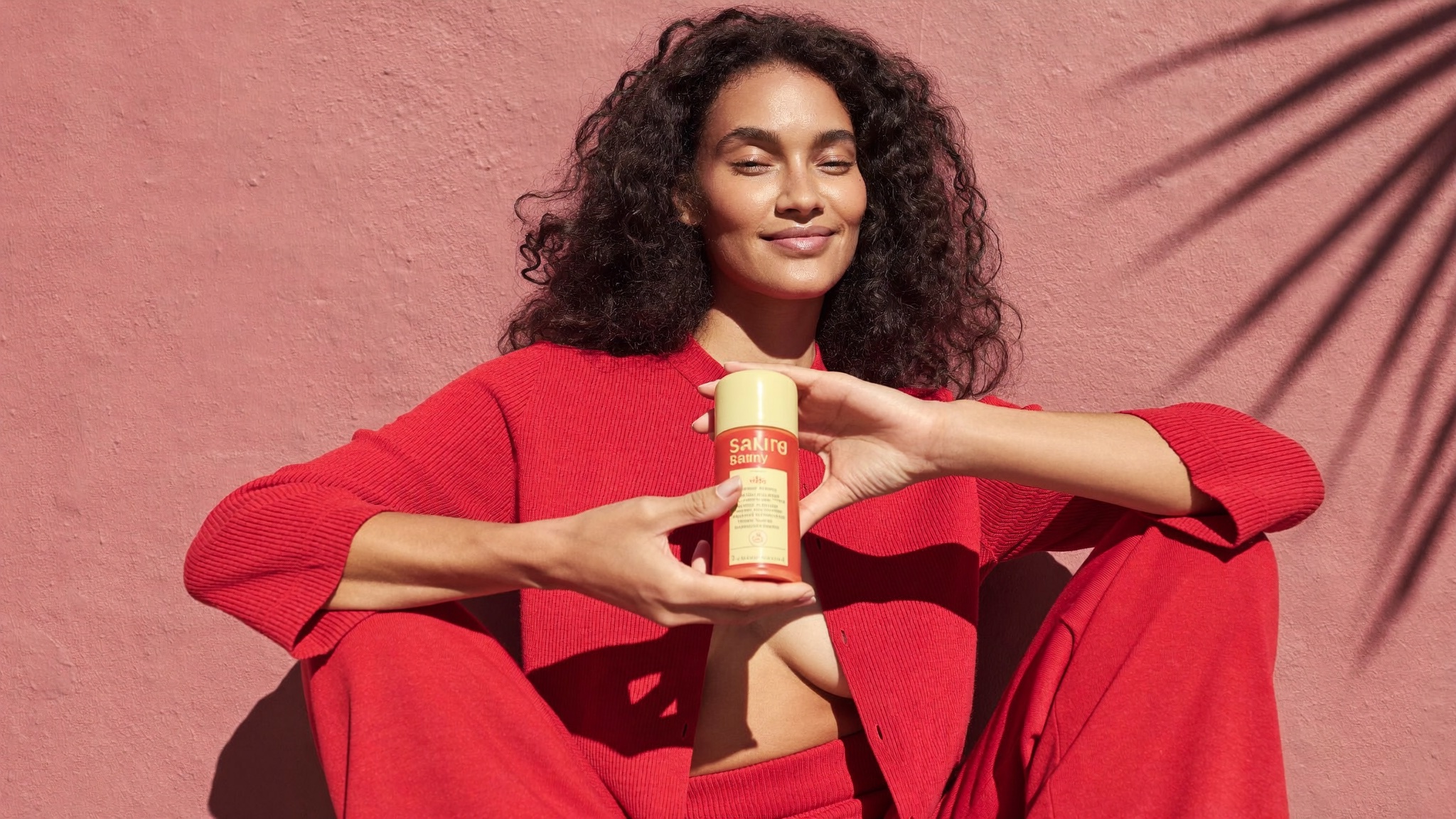A creator in red presenting an orange bottle against a pink wall.