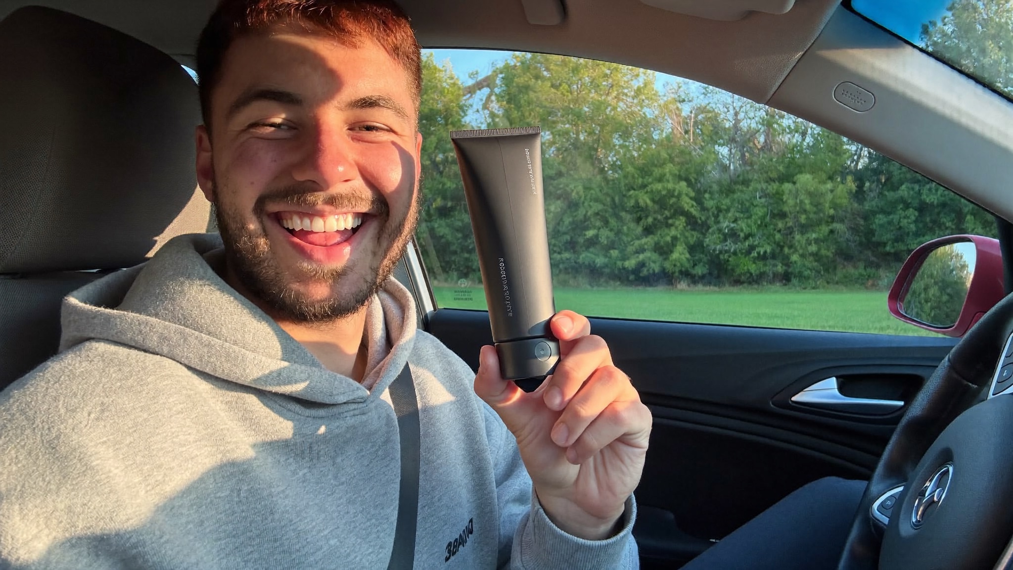 A smiling creator holding a black product tube in a sunlit car.